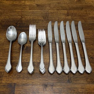 Puede incluir: Una colección de cubiertos de color plateado, que incluye cucharas, tenedores y cuchillos, dispuestos sobre una superficie de madera. Las cucharas y tenedores tienen un diseño decorativo con volantes en la base de los mangos. Los cuchillos tienen una forma simple y elegante.