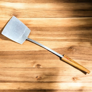 May include: A stainless steel spatula with a rectangular head and a long, thin handle. The handle has a wooden grip. The spatula is set against a wooden background.