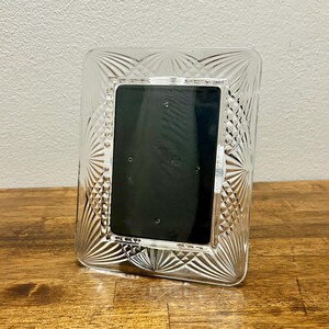 May include: A clear glass picture frame featuring a rectangular center opening and a wide border decorated with a pressed fan-like pattern and diamond-shaped cross-hatching. The frame has a black insert and a smooth, reflective surface finish.