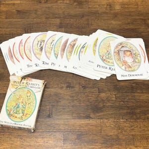 May include: A Peter Rabbit's Giant Picture Card Game set featuring a rectangular cardboard box with an illustration of a rabbit and a bird, alongside a fan of playing cards with colorful borders and character names like Miss Dormouse and Peter Rabbit.