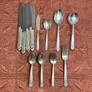 May include: A collection of vintage silverware, including knives, spoons, forks, and a butter knife, arranged on a patterned, reddish-brown surface. The silverware has ornate floral detailing on the handles and a shiny, reflective finish.