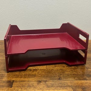 Könnte beinhalten: Ein dunkelroter Schreibtisch-Organizer aus Kunststoff mit zwei übereinander gestapelten Ablagefächern. Das Gerät verfügt über ein offenes Design mit seitlichen Aussparungen für den einfachen Zugriff auf Dokumente. Die Struktur ist modular.