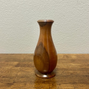 May include: A small, handcrafted wooden vase with a smooth, polished finish. The vase has a rounded body and a flared top, showcasing the natural wood grain in shades of brown and tan. It stands on a small, circular base, perfect for displaying small flowers or decorative items.