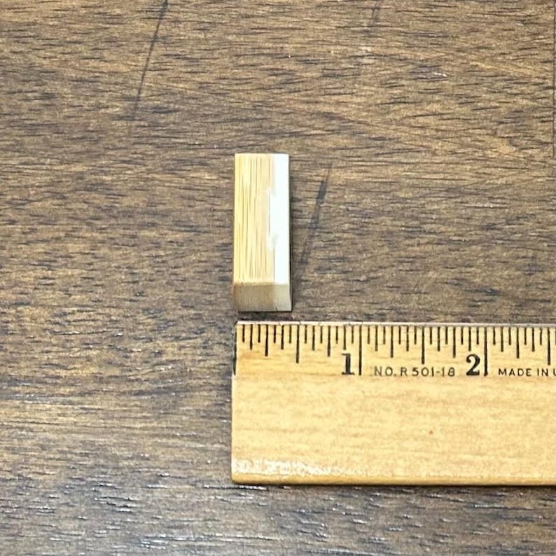 May include: A small, rectangular wooden block with a light brown and white striped pattern. The block is next to a wooden ruler, showing its length is approximately 0.75 inches.
