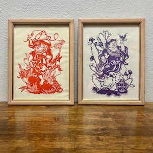 May include: Two framed paper cutouts in light wood frames. The left frame contains a red paper cutout of a figure holding coins. The right frame contains a purple paper cutout of a figure holding a bat, standing next to a cactus and a round vessel.