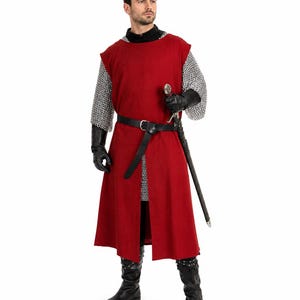 Puede incluir: Una túnica roja sin mangas sobre una camisa de cota de malla plateada de manga larga. El conjunto incluye un cinturón de cuero negro con hebilla plateada, guantes de cuero negro y botas de cuero negro. Se sostiene una espada con empuñadura plateada.
