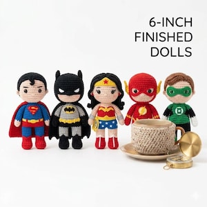 May include: Five 6-inch finished crochet dolls representing superhero characters including Superman, Batman, Wonder Woman, The Flash, and Green Lantern, standing next to a beige crocheted mug cozy on a saucer and a small gold compass.