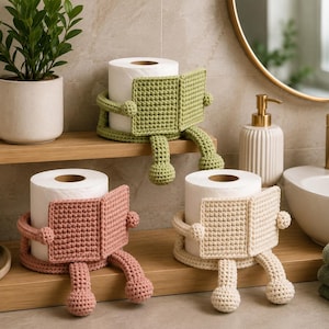 May include: Three crochet toilet paper holders in sage green, dusty rose, and cream. Each holder features a character design with arms holding a square panel and two dangling legs with rounded feet, designed to wrap around a standard toilet paper roll.