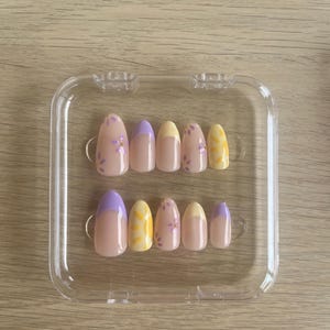 May include: A set of ten press-on nails in a clear plastic case. The nails feature a nude base with various designs including lavender French tips, yellow sun motifs, and small purple floral patterns with tiny gold accents on an almond-shaped base.