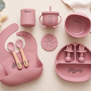 May include: A dusty rose silicone feeding set including a bib, a cup with a straw and giraffe graphic, a bowl, a snack lid, a divided plate, and utensils with wooden handles. The name Hayley is printed on the bib, cup, and plate in a dark font.