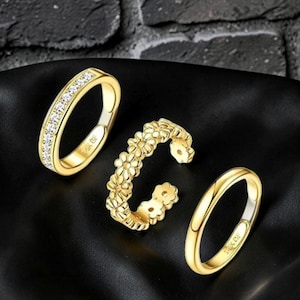 Puede incluir: Un conjunto de tres anillos dorados. Un anillo presenta una hilera de piedras transparentes, otro es una banda abierta con un patrón repetido de flores tipo margarita y el tercero es una banda dorada lisa. Cada anillo tiene la inscripción 18K GF.