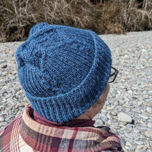 May include: A blue knitted beanie hat with a textured design. The hat appears to be made of wool. The person is wearing a plaid shirt in shades of red, brown, and beige.