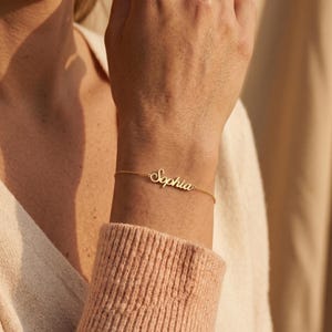 Könnte beinhalten: Ein goldfarbenes Metallkettenarmband mit einer zentralen Namensplatte, auf der der Name Sophia in Schreibschrift eingraviert ist. Die feine Kette ist beidseitig mit der Namensplatte verbunden und bildet ein durchgehendes Schmuckstück.