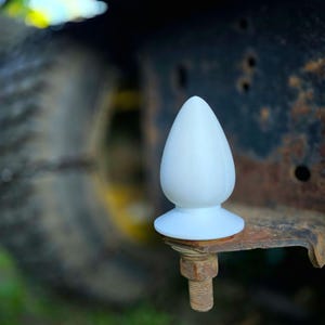 May include: A smooth, opaque white, teardrop-shaped object with a flared base, mounted onto a threaded metal bolt that is attached to a rusted, dark-colored metal bracket, positioned in front of a blurred, dark, heavy-duty tire.