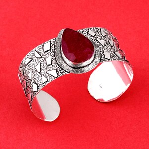 Può includere: Un bracciale a fascia in argento con una pietra preziosa rosso scuro a forma di goccia al centro. La fascia presenta una finitura ossidata con un motivo strutturato di piccole forme a foglia e accenti geometrici triangolari lucidi.