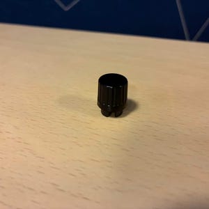 May include: A small, cylindrical black control knob featuring a smooth, flat top surface and a vertically ribbed side texture. The base of the knob includes a small notch or cutout, indicating a specific mounting orientation for electronic equipment.
