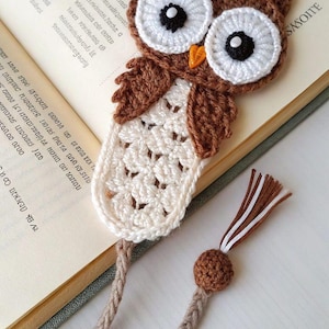 May include: A crocheted owl bookmark featuring a brown head with large white and black eyes, a small orange beak, and a cream-colored body with a textured pattern. Two braided brown cords extend from the bottom, ending in a small brown bead and a tassel.