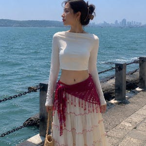 May include: A long-sleeved white crop top paired with a tiered cream-colored maxi skirt featuring red trim. A dark magenta crocheted hip scarf with dangling fringe accents is tied at the waist, accompanied by a woven straw tote bag.