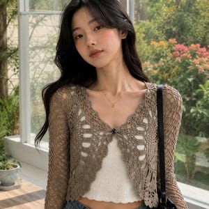 May include: A long-sleeved, taupe-colored crochet cardigan featuring an open-knit, web-like pattern with a scalloped edge. The cardigan has a single dark button closure at the center of the V-neckline and is worn over a white, sleeveless top.