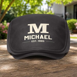 Peut inclure: Un coussin appuie-tête en cuir texturé gris anthracite foncé arborant une grande lettre M de couleur crème sur le dessus, avec le nom MICHAEL imprimé en dessous, suivi du texte EST. 1985 centré entre deux fines lignes horizontales.