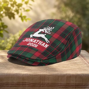 May include: A flat cap featuring a red, green, and black plaid pattern. The front displays a white graphic of a leaping reindeer above a red banner containing the white text JONATHAN 2026. The cap has a structured, rounded crown and a short brim.