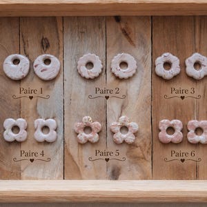 Peut inclure: Six paires de boucles d'oreilles circulaires et en forme de fleur dans des tons blanc, beige et rose avec une texture marbrée. Chaque paire est étiquetée avec le texte de Paire 1 à Paire 6, avec une petite icône de cœur sous chaque étiquette.