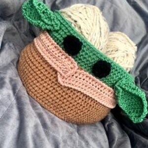May include: A crocheted storage basket shaped like a green character with large ears, black eyes, and a pink mouth. The basket is made with brown, pink, and green yarn.