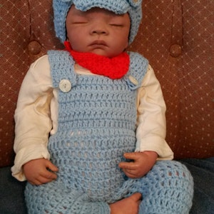 May include: A baby doll wearing a light blue crocheted outfit, including overalls and a cap with button details. The doll also has a red bib and a white long-sleeved shirt. The doll is sitting on a blue surface.