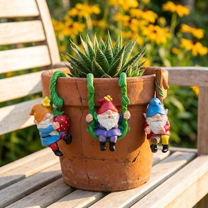 May include: A terracotta flower pot containing a green succulent plant, featuring three small gnome figurines attached to the rim. One gnome holds a red strawberry, one sits on a green vine swing, and one sits on a small wooden log.