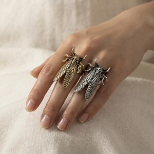 Può includere: Due anelli a forma di cicala, uno con finitura in oro metallizzato e l'altro con finitura in argento metallizzato. Ogni anello presenta ali e zampe dettagliate, con il corpo dell'insetto che si estende lungo il dito.
