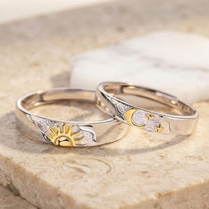 Könnte beinhalten: Zwei silberfarbene Ringe mit strukturierter Oberfläche. Ein Ring zeigt ein goldfarbenes Sonnenmotiv, während der andere Ring ein goldfarbenes Mondsichel- und Sterndesign aufweist. Beide Ringe haben polierte silberne Kanten.