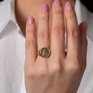以下が含まれることがあります： 中央に文字「A」が刻印された円形のフェイスを持つゴールドのシグネットリング。リングは光沢のある仕上げで、指にフィットする滑らかで丸みを帯びたバンドが特徴です。デザインはシンプルで、文字のディテールに焦点を当てています。