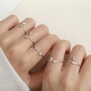 May include: A collection of silver-toned open rings worn on fingers, each featuring a single letter charm encrusted with small clear stones. The letters visible include V, W, X, and L, with each ring band also accented by a small clear stone.