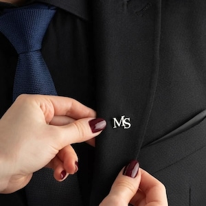 May include: A silver-toned metal lapel pin featuring the letters MS in a serif font, attached to the lapel of a black blazer. The blazer is worn over a black shirt and a textured navy blue necktie, creating a formal and professional appearance.
