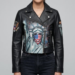 Op de afbeelding: Een zwart leren motorjack met een grote grafische print van het Vrijheidsbeeld met een zonnebril en een bandana met Amerikaanse vlag. De jas is voorzien van een adelaar-patch op de rechtermouw en een fakkel-patch op de linkermouw.