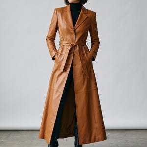 May include: A long, tan leather trench coat featuring a notched lapel, a button-front closure, and a matching tie belt at the waist. The coat has long sleeves, structured shoulders, and a floor-length hemline, worn over a black turtleneck and trousers.