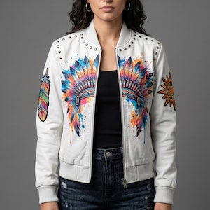 May include: A white bomber jacket featuring a colourful watercolour-style graphic of a feathered headdress on the chest, a single feather on the right sleeve, and a sun emblem on the left sleeve, accented with silver studs along the shoulder seams.
