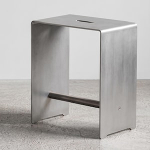 Könnte beinhalten: Ein rechteckiger Hocker aus gebürstetem, silberfarbenem Metall. Das Design besteht aus einer flachen Sitzfläche mit einem mittigen, ovalen Griffausschnitt und zwei vertikalen Seitenteilen, die durch eine zylindrische Stange verbunden sind.