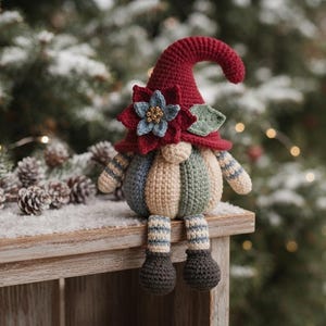May include: A crocheted gnome figurine featuring a tall, dark red pointed hat adorned with a blue and red flower accent. The gnome has a round, beige body with vertical stripes in muted blue, green, and cream, and dangling striped legs with dark feet.