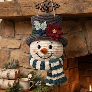 Pu&ograve; includere: Un ornamento a forma di pupazzo di neve all'uncinetto con un cappello a cilindro grigio scuro decorato con una stella di Natale rossa, foglie verdi e bacche blu. Il pupazzo ha la testa bianca, un naso arancione e una sciarpa a righe.