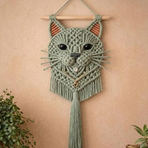May include: A sage green macrame wall hanging shaped like a cat head, featuring dark eyes, a small nose, and whiskers. The design includes inner ear details in a lighter shade, a gold-toned chain necklace with a heart charm, and a long fringe tassel.