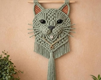 Può includere: Una decorazione da parete in macramè color verde salvia a forma di testa di gatto, con occhi scuri, un piccolo naso e baffi. Il design include dettagli più chiari all'interno delle orecchie, una catenina dorata con un cuore e una lunga frangia.