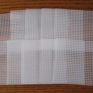 May include: A stack of white plastic canvas sheets with a grid pattern. The sheets are rectangular and vary slightly in size, with a few overlapping. The material appears to be translucent, and the sheets are arranged on a wooden surface.