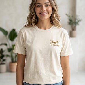 May include: A cream-coloured short-sleeve t-shirt featuring the text "Angel in Training" printed in a brown script font on the upper left chest area, accompanied by a small graphic of two wings positioned above the word Angel.