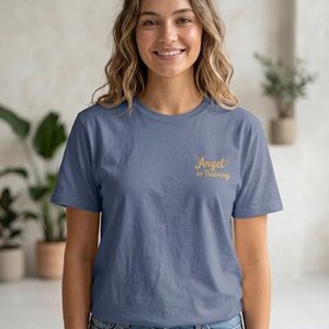 May include: A slate blue short-sleeved t-shirt featuring the text "Angel in Training" printed in a gold script font on the upper left chest area, accompanied by a small graphic of two wings positioned above the word Angel.