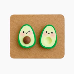Può includere: Un paio di orecchini a lobo a forma di metà avocado con buccia verde, polpa verde chiaro e guance rosa. Un orecchino presenta un nocciolo marrone, mentre l'altro ha un centro cavo. Gli orecchini sono fissati su un cartoncino marrone.