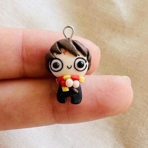 May include: A small, handcrafted polymer clay charm featuring a stylized character with dark brown hair, large black eyes, and a lightning bolt mark on the forehead. The figure wears a black outfit and a red and yellow striped scarf with a metal loop.