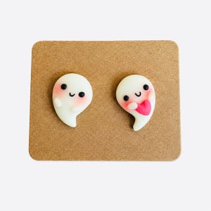Può includere: Un paio di orecchini a lobo a forma di piccoli fantasmi bianchi con occhi neri, bocche sorridenti e guance rosa. Un fantasma ha le braccia sollevate, mentre l'altro tiene un piccolo cuore rosa contro il petto, fissati su un cartoncino marrone.