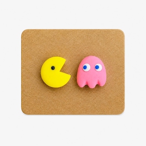 Peut inclure: Un personnage circulaire jaune avec une bouche en forme de coin et un œil noir, placé à côté d'un personnage rose en forme de fantôme avec deux grands yeux blancs à pupilles bleues, sur une surface rectangulaire brune texturée.