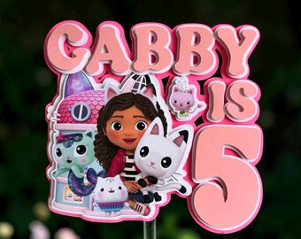 May include: A layered paper cake topper featuring the text GABBY IS 5 in pink and white. The design includes a cartoon character with brown hair and cat ears, a small pink cat, a white cat, a teal mermaid cat, and a pink building structure.